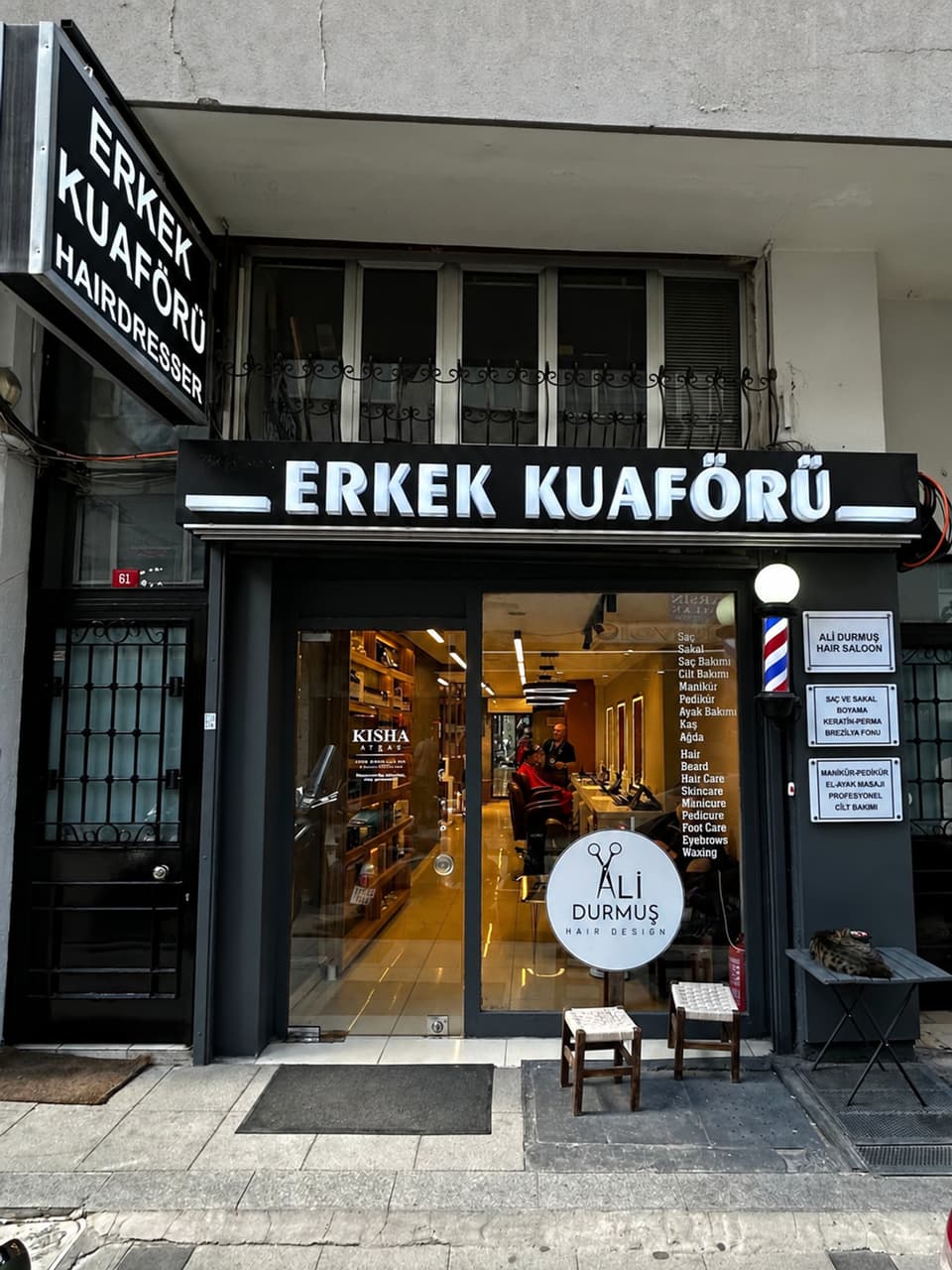 Koca Mansur Sokak 61'deki dükkanın vitrini: Erkek Kuaförü tabelası, berber direği ve kapıdaki hizmet levhaları